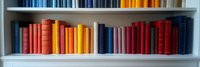 Colorful hardcover books arranged by color spectrum on white bookshelf, creating rainbow gradient effect from blue through yellow to red spines. Library organization concept.