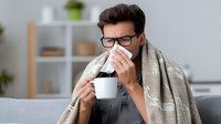 Young man with glasses and blanket blowing nose while holding hot drink on couch at home, suffering from cold or flu symptoms during illness.