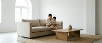 Young Asian mother and toddler son bonding on beige sofa in minimalist living room with natural light from arched window and wooden coffee table.