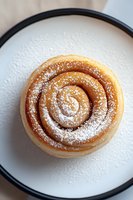 Freshly baked cinnamon roll dusted with powdered sugar on white plate with black rim, viewed from above. Close-up shows spiral pattern and golden pastry.