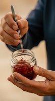 Close-up of hands holding glass jar with red jam or preserve, stirring with metal spoon. Suitable for food, cooking, and culinary content.