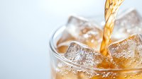 Pouring golden carbonated soda drink into glass with ice cubes, close up view of refreshing beverage with bubbles and splashes on light background.