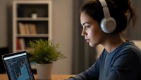Young woman wearing white headphones working on laptop computer at home office desk with plant in background during evening study session.