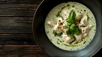 Creamy chicken soup with fresh basil and black pepper served in dark ceramic bowl on rustic wooden table, overhead view with copy space.