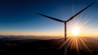 Wind turbine silhouetted against dramatic sunset sky with sun rays bursting through blades, mountain range in background creates scenic renewable energy landscape.
