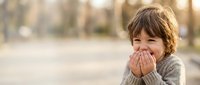 Happy young boy with brown hair covering mouth while laughing outdoors in warm sunlight during golden hour creating joyful childhood moment.
