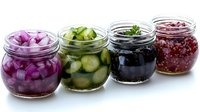 Fresh pickled vegetables and berries in glass jars. Homemade preserves with red onion, cucumber, olives and raspberry jam arranged in row on white surface.