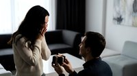 Young Caucasian man proposing to surprised Asian woman with engagement ring in modern living room. Romantic marriage proposal moment at home.