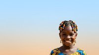 Young African girl with colorful braided hairstyle and beaded hair accessories smiling outdoors against clear sky backdrop. Portrait suitable for diversity, childhood, and cultural representation projects.