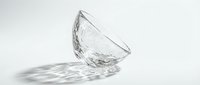 Tilted clear glass bowl with textured surface casting intricate light shadows on white background, minimalist elegant tableware concept.