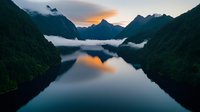Tranquil fjord landscape at dawn with low-lying clouds between mountain peaks, dramatic sky reflection in still waters creates mirror effect in Nordic scenery.