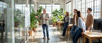 Group of diverse young professionals collaborating in bright modern office space with plants, discussing projects and sharing ideas in contemporary workplace environment.