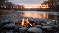 Cozy campfire burning bright on rocky riverbank at golden sunset with peaceful water reflections and bare winter trees creating warm outdoor adventure atmosphere.
