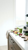 Green smoothie in a glass on a bright kitchen counter with fresh berries, banana slices, spinach leaves, a bowl, cutting board, and blender nearby.