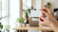 Hands holding blank white business card in modern office workspace with plants and natural light.