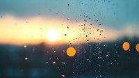 Raindrops on window glass during sunset with blurred bokeh lights creating atmospheric mood. Water droplets catch golden hour sunlight against darkening sky.