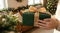Hands holding green gift box with gold ribbon bow near Christmas tree with warm lights creating festive holiday atmosphere for seasonal celebrations.