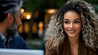 Young woman with curly hair and warm smile meets man in business casual attire outdoors at dusk. Evening lighting creates romantic atmosphere for dating, relationship, or professional networking concepts.