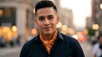 Young Hispanic man with stylish haircut wearing black jacket and orange patterned shirt smiling confidently on urban street with warm bokeh lights background.