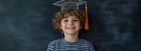 Young student wearing graduation cap and striped shirt smiles brightly against dark chalkboard background, expressing joy and achievement in education.