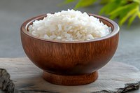 Steamed white jasmine rice served in wooden bowl on stone surface with green plants in background, close up view of traditional Asian cuisine.