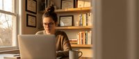 Woman with brown hair and glasses wearing a tan cardigan working on laptop at desk by window with bookshelves, warm lighting, professional home office setting.