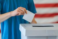 Person in blue t-shirt casting ballot at polling station with American flag in background, perfect for election coverage, democracy and voting rights concepts.