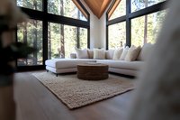Modern living room with large windows overlooking forest, white sectional sofa, woven coffee table, and textured area rug in contemporary A-frame interior design.