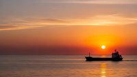 Cargo ship silhouette sailing on calm ocean waters during golden sunset with warm orange sky reflecting on sea surface for maritime transport concepts.