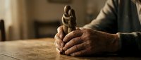 Senior man with weathered hands holding small wooden figurine on table, representing craftsmanship, nostalgia, and handmade artisan work for heritage and tradition themes.