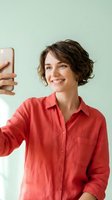 Happy young woman with short curly brown hair taking selfie with smartphone wearing coral red shirt against mint green background for social media content.