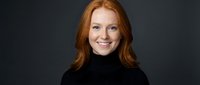 Professional redhead businesswoman in black turtleneck smiling confidently against dark background for corporate headshot and marketing materials.