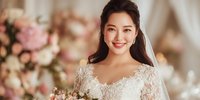 Young Asian bride in elegant white lace wedding dress smiling warmly at camera with soft romantic lighting and pink floral arrangements in luxurious bridal suite.