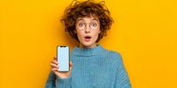 Surprised young woman with curly hair and glasses holding smartphone with blank screen against bright yellow background for mobile app mockup.