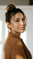 Young Hispanic woman with messy bun hairstyle wearing brown turtleneck sweater and gold hoop earrings looking over shoulder with confident smile in natural light.