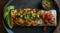 Grilled burrito filled with melted cheese and topped with fresh avocado slices, diced tomatoes and cilantro, served with spicy salsa verde on rustic plate.