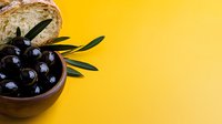 Black olives in ceramic bowl with fresh bread and olive branch on bright yellow background, food styling for culinary and recipe content.