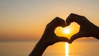 Hands forming heart shape silhouette against golden sunset over calm water creating romantic atmosphere for love and connection concepts.