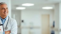 Smiling senior male doctor with stethoscope in white coat standing confidently in modern hospital corridor with arms crossed looking at camera.