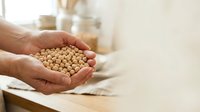Adult hands holding dried chickpeas in kitchen setting with blurred storage containers background. Suitable for food, nutrition, cooking, and healthy eating content.