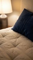 Cozy bedroom corner with navy blue pillow on beige quilted bedding, warm table lamp with metallic base, and neutral textured wallpaper creating inviting sleeping space.