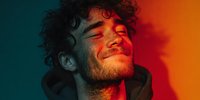 Young Caucasian man with curly hair and stubble smiling with eyes closed in dramatic lighting with orange and teal gradient background.