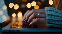 Female hands typing on laptop keyboard in evening with warm bokeh lights in background, cozy sweater sleeves visible, creating atmospheric home office mood.