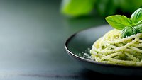 Spaghetti pasta with fresh basil pesto sauce and parmesan cheese served in dark ceramic bowl on black background. Fresh herbs garnish. Food photography.