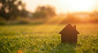 Small wooden house model on green grass field during golden hour sunset with warm bokeh background for real estate and home concepts.