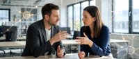 Young professional man and woman in business casual attire discussing project ideas at modern office desk with coffee cups, bright windows and city skyline visible behind them.