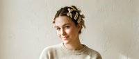 Young Caucasian woman with braided updo adorned with bow hair accessories, wearing a cozy beige sweater, smiling softly against a neutral textured wall.