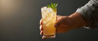 Man's hand holding refreshing golden cocktail with ice and mint garnish against dark background, perfect for beverage and hospitality marketing.