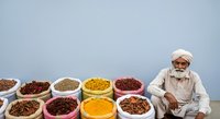 Elderly male vendor of South Asian ethnicity wearing traditional white turban and clothing, seated beside colorful bowls and containers filled with various dried spices, grains, and legumes in warm earth tones against blue background for commerce or food trade concepts.