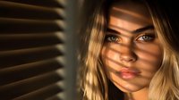 Young blonde woman with intense gaze in dramatic lighting through window blinds creating shadow stripes across face, moody atmospheric portrait in warm golden tones.